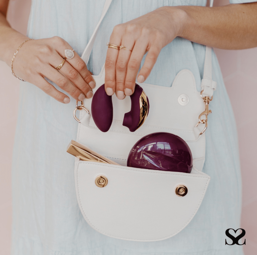 A woman's torso pictured and she is wearing a pale blue dress and has a white handbag at her hip which is open and showing a purple small round shiny item in her bag that looks like it contains makeup.